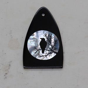 May include: A black, pointed, shield-shaped pendant featuring a circular inlay of iridescent mother-of-pearl. The inlay displays a black silhouette of a bird perched on a bare tree branch. A small circular hole is positioned at the top center.