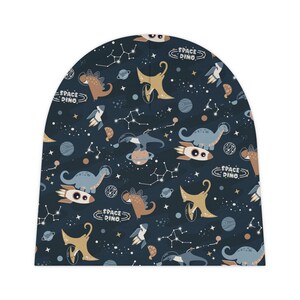 May include: A blue beanie with a repeating pattern of dinosaurs riding rockets in space. The pattern also includes stars, planets, and the text "Space Dino".