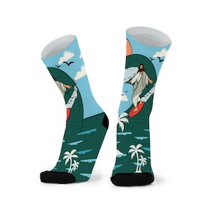 May include: A pair of socks with a cartoon illustration of Jesus surfing a wave. The socks have a blue sky background with clouds, birds, and a setting sun. Palm trees and ocean waves complete the design.