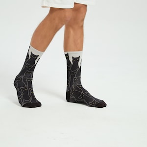 May include: Black socks featuring a repeating pattern of cat silhouettes. The socks have a light grey cuff and a black foot. The image shows the socks being worn on a person's legs.