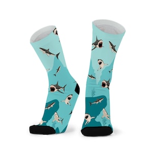 Könnte beinhalten: Ein Paar Crew-Socken mit einem sich wiederholenden Muster von Haien in verschiedenen Posen vor einem hellblauen Hintergrund. Die Haie sind in Grautönen, Weiß und Schwarz dargestellt, mit geöffneten Mäulern. Die Socken haben schwarze Fersen und Zehen.