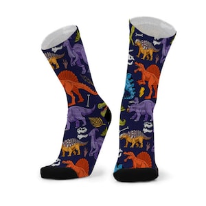 以下が含まれることがあります： 恐竜柄の青い靴下2足組。オレンジ、パープル、グリーンなど、さまざまな色の恐竜が描かれています。恐竜の周りは骨と葉で囲まれています。