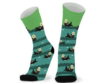 May include: A pair of crew socks featuring a repeating pattern of brown sea otters floating on their backs. The background consists of horizontal wavy stripes in shades of teal and green, with solid black toe and heel sections.