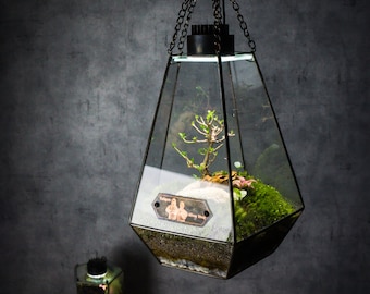 May include: A hanging glass terrarium with a geometric shape, featuring a small plant, moss, and a decorative plaque with the text "Together we grow" in a cursive font. The terrarium is illuminated from within, casting a warm glow.