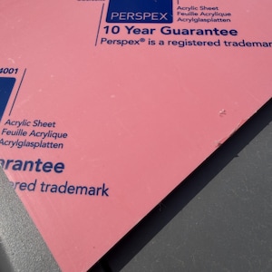 May include: A flat, opaque pink acrylic sheet with blue printed text. The text includes the brand name Perspex, the phrase 10 Year Guarantee, and the product description Acrylic Sheet, Feuille Acrylique, Acrylglasplatten, and the number 4001.