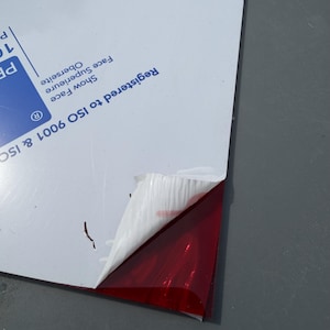 May include: A rectangular sheet of translucent red acrylic material with a white protective film partially peeled back from one corner. The white film features blue printed text that reads Registered to ISO 9001 & ISO 14001.