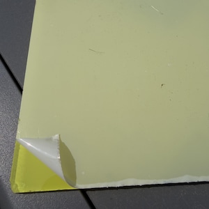 May include: A rectangular sheet of pale yellow material with a smooth, flat surface. One corner of the sheet features a white adhesive backing that is partially peeled back, revealing the bright yellow underside of the material beneath the layer.