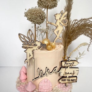 May include: A light pink cake decorated with wooden fairy cutouts, a sign that says "The way to Luna's Fairy Tea Party", and dried flowers.