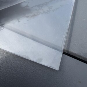 May include: A thin, rectangular sheet of clear, transparent plastic material with a smooth surface. The sheet is partially lifted at one corner, revealing a slight flexibility and a glossy finish that reflects light across its flat, uniform surface.