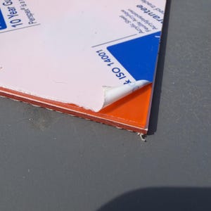 May include: A rectangular sheet of orange acrylic material with a protective white paper film partially peeled back from one corner. The white film features printed text including 10 Year Guarantee, Perspex, and ISO 14001 branding details.