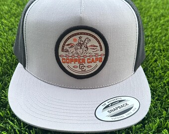 May include: A gray and black trucker hat with a curved brim. The hat features a circular patch with a cowboy riding a horse, the words "Copper Caps," and the letter "G."