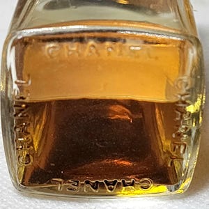 May include: A small, rectangular glass bottle filled with a golden-colored liquid. The word "CHANEL" is embossed on the front and sides of the bottle. The bottle has a clear, transparent appearance.