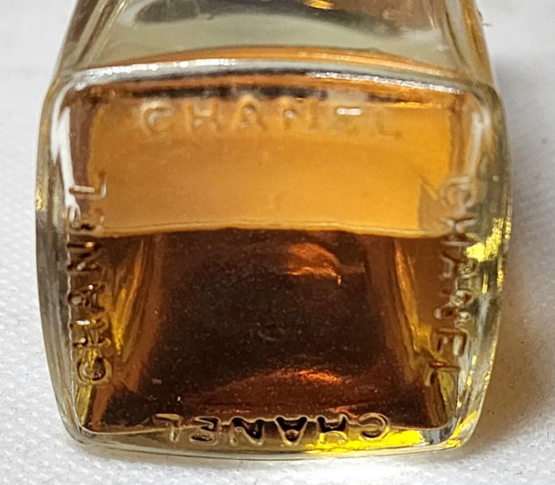 May include: A small, rectangular glass bottle filled with a golden-colored liquid. The word "CHANEL" is embossed on the front and sides of the bottle. The bottle has a clear, transparent appearance.