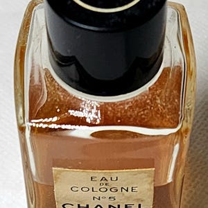 May include: A small, rectangular perfume bottle of Chanel No. 5 Eau de Cologne. The bottle is filled with a light brown liquid and has a black cap with the Chanel logo. The label reads "Chanel Paris".