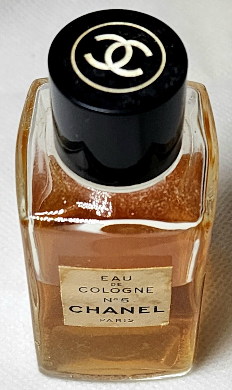 May include: A small, rectangular perfume bottle of Chanel No. 5 Eau de Cologne. The bottle is filled with a light brown liquid and has a black cap with the Chanel logo. The label reads "Chanel Paris".