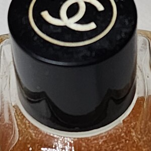 May include: Close-up of a perfume bottle with a black cap. The cap features a white double-C logo within a white circle. The bottle's contents are a light brown color, visible through the clear glass.