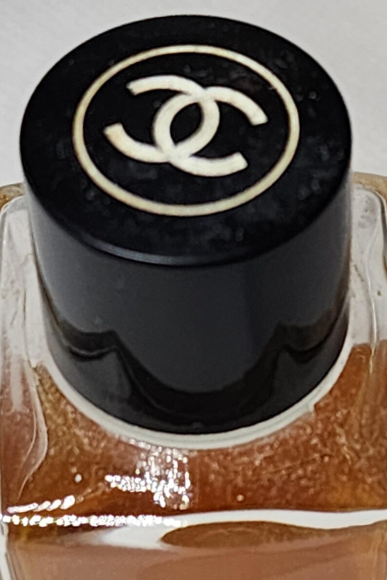 May include: Close-up of a perfume bottle with a black cap. The cap features a white double-C logo within a white circle. The bottle's contents are a light brown color, visible through the clear glass.