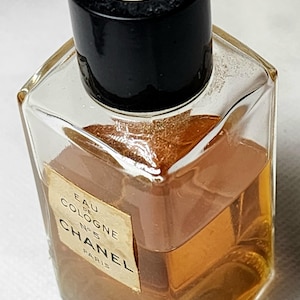 May include: A vintage Chanel No. 5 Eau de Cologne bottle. The clear, square-shaped glass bottle contains a golden-brown liquid. The black cap features a white interlocking CC logo. The label reads "Chanel Paris".