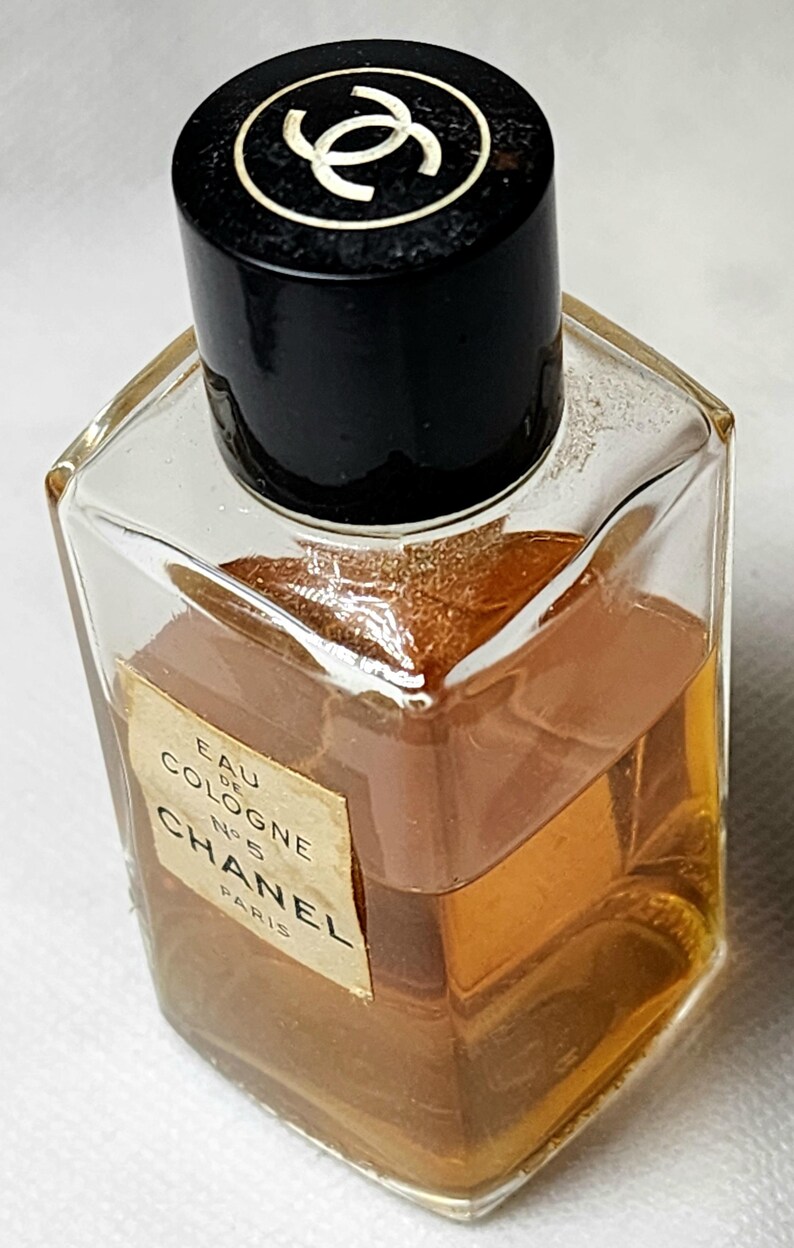 May include: A vintage Chanel No. 5 Eau de Cologne bottle. The clear, square-shaped glass bottle contains a golden-brown liquid. The black cap features a white interlocking CC logo. The label reads "Chanel Paris".