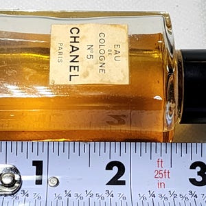 May include: A small, rectangular Chanel No. 5 Eau de Cologne bottle with a black cap. The bottle contains a golden-colored liquid. The label reads "CHANEL PARIS N&deg;5 EAU DE COLOGNE". The bottle is approximately 3 inches long.