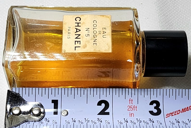 May include: A small, rectangular Chanel No. 5 Eau de Cologne bottle with a black cap. The bottle contains a golden-colored liquid. The label reads "CHANEL PARIS N&deg;5 EAU DE COLOGNE". The bottle is approximately 3 inches long.