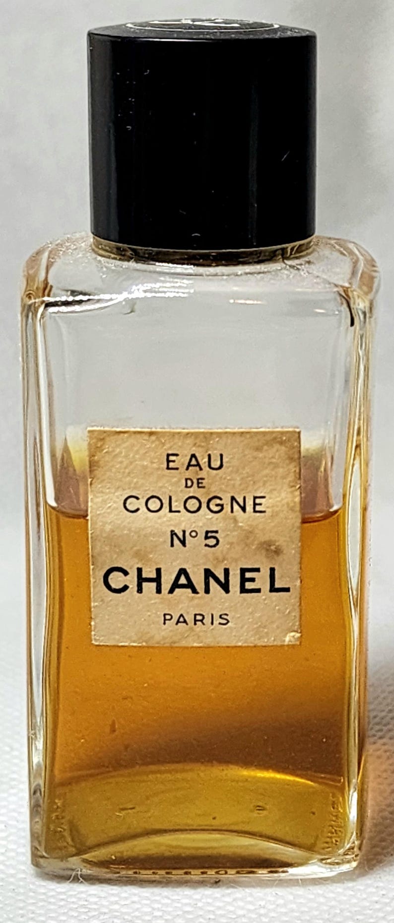 May include: A vintage Chanel No. 5 Eau de Cologne bottle. The clear glass bottle contains a golden liquid and has a black cap. The label reads "Eau de Cologne No. 5 Chanel Paris."
