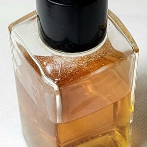 May include: A small, clear glass perfume bottle with a black cap. The bottle contains a light amber liquid. The cap features a white logo on a black background. The bottle has a square shape.