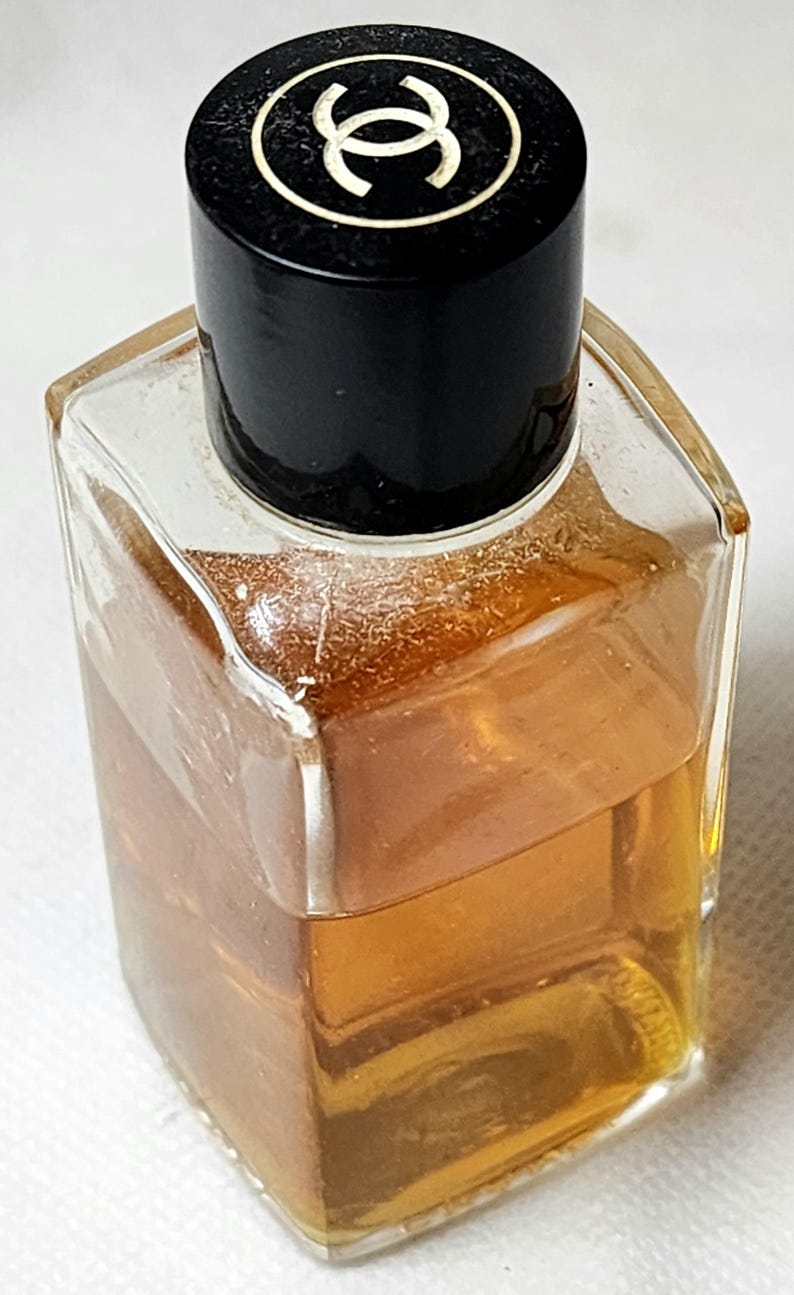 May include: A small, clear glass perfume bottle with a black cap. The bottle contains a light amber liquid. The cap features a white logo on a black background. The bottle has a square shape.