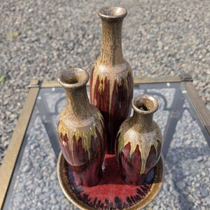 May include: A set of three ceramic vases of varying heights arranged on a matching circular tray. The vases and tray feature a deep red base color with a tan and yellow drip glaze effect cascading from the narrow necks down the bodies.