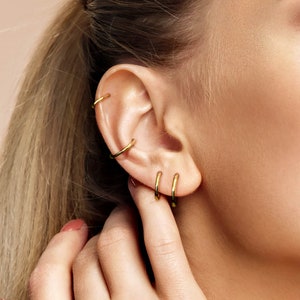 May include: Four thin, polished gold-tone hoop earrings worn in a single ear. Two hoops are positioned along the upper cartilage, while two smaller hoops are placed in the lower earlobe. The jewelry features a smooth, circular metal design.