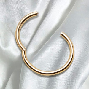 May include: A circular gold hoop earring with a hinged segment closure. The metal surface is smooth and polished, reflecting light. The text 14K is clearly engraved on the side of the hoop near the hinge, indicating the gold purity of the piece.