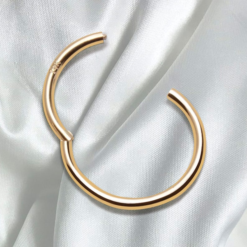 May include: A circular gold hoop earring with a hinged segment closure. The metal surface is smooth and polished, reflecting light. The text 14K is clearly engraved on the side of the hoop near the hinge, indicating the gold purity of the piece.