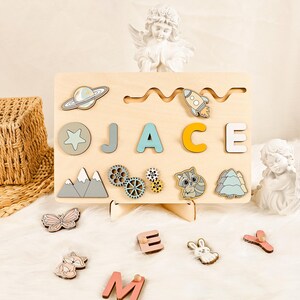 Puede incluir: Puzzle de madera con el nombre "JACE" en letras azules, amarillas y blancas. El rompecabezas también incluye un cohete, un planeta, una estrella, montañas, engranajes, un mapache y un pino.