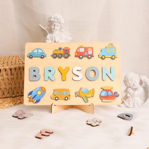 May include: Wooden puzzle featuring colourful vehicles and the name BRYSON in large letters. The puzzle pieces are shaped like the letters and vehicles. The puzzle is on a wooden stand.