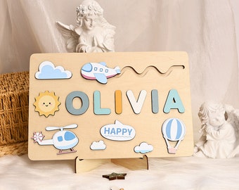 May include: Wooden puzzle with a light brown background featuring the name "OLIVIA" in colorful letters. The puzzle includes a blue airplane, a helicopter, a hot air balloon, a sun, and clouds. The puzzle also includes a speech bubble with the word "HAPPY" on it.