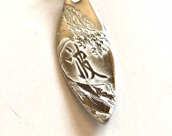 May include: A metallic pendant with a detailed design. The pendant is a light gold color, with a textured surface and a stylized image of a person. The pendant hangs from a thin, dark chain.