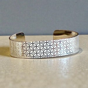 May include: A silver cuff bracelet with a repeating geometric pattern. The open cuff design features a series of interlocking circles and squares, creating a textured surface. The bracelet is made of a shiny, reflective metal.