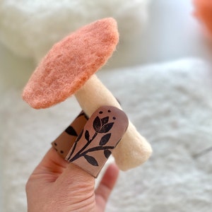 Puede incluir: Un hongo de fieltro con un sombrero de color melocotón y un tallo blanco. El tallo está envuelto en un títere de dedo de cuero marrón con un diseño floral negro.