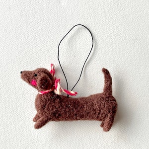 May include: A brown felt dachshund ornament with a pink and white striped ribbon around its neck. The ornament has a black string for hanging.