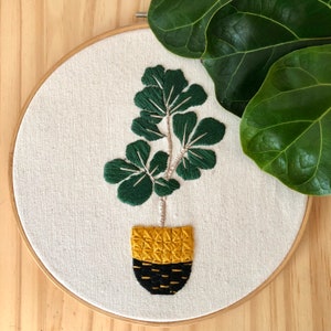 Peut inclure: Art de broderie sur un cercle représentant un ficus vert dans un pot jaune et noir. La plante est brodée avec des détails et le pot a un motif géométrique.
