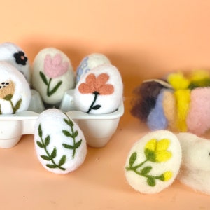 Peut inclure: Six œufs de Pâques en feutre blanc avec différents motifs floraux brodés dans un carton à œufs blanc. Deux autres œufs en feutre avec des broderies florales sont sur une table avec une aiguille et un tas de laine colorée.