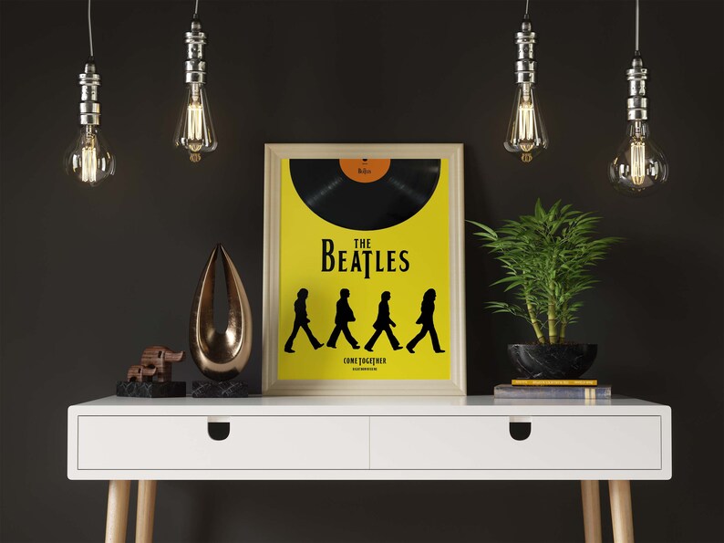 Puede incluir: Un escritorio blanco con dos cajones y un p&oacute;ster enmarcado de The Beatles con el texto "The Beatles Come Together" sobre un fondo amarillo. El p&oacute;ster presenta una silueta negra de los miembros de la banda caminando por una calle. Hay tres bombillas colgantes sobre el escritorio.