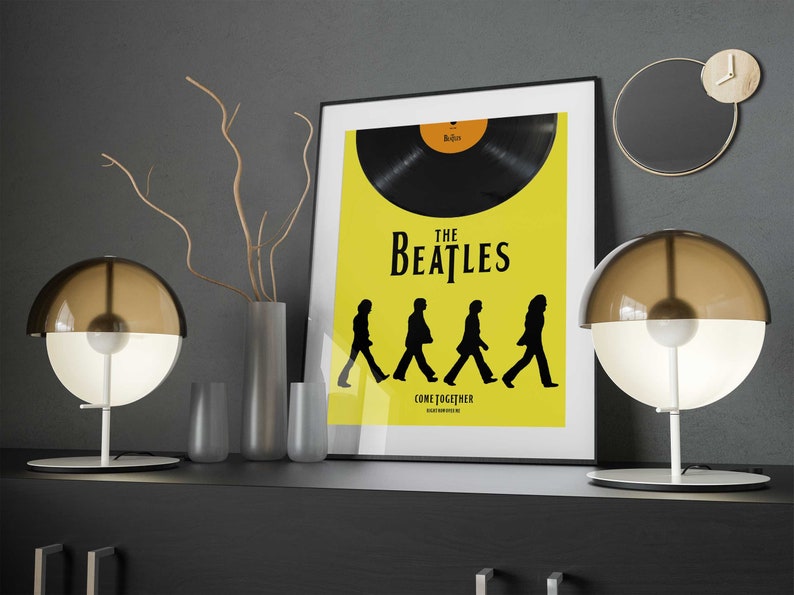 Puede incluir: Un p&oacute;ster enmarcado en negro con fondo amarillo que presenta un disco de vinilo negro y el texto "The Beatles" y "Come Together". El p&oacute;ster se exhibe en un estante con dos l&aacute;mparas de globo marr&oacute;n y blanco y un jarr&oacute;n con ramas secas.