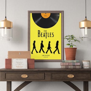 Puede incluir: Un p&oacute;ster enmarcado con fondo amarillo que presenta una silueta negra de los Beatles caminando por el p&oacute;ster. El p&oacute;ster tiene el texto "The Beatles" y "Come Together" impreso en &eacute;l. El p&oacute;ster est&aacute; colgado en una pared blanca sobre una mesa de madera con dos cajones. Hay dos l&aacute;mparas colgantes doradas colgadas sobre la mesa, y una planta en maceta est&aacute; sobre la mesa a la derecha del p&oacute;ster.