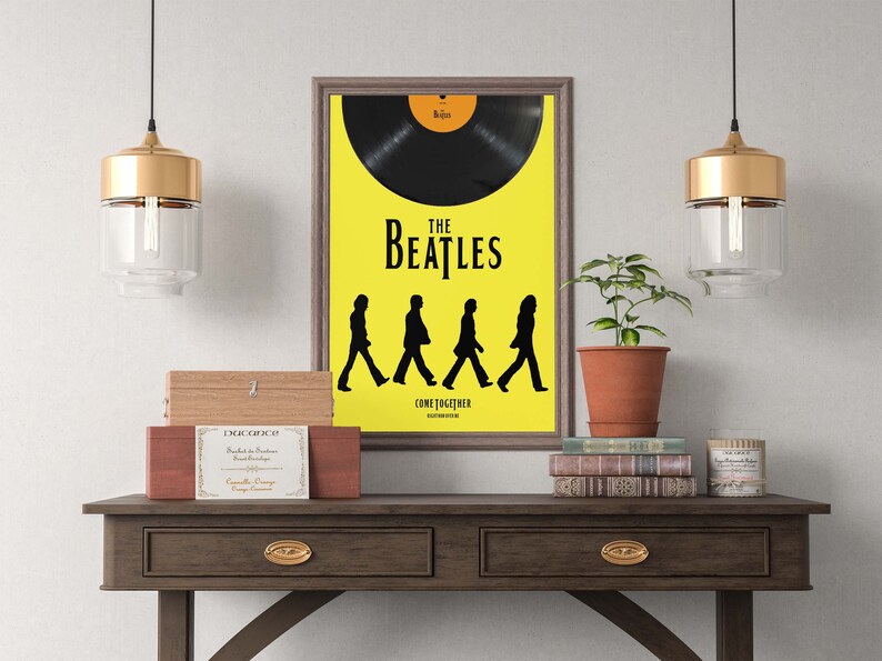 Puede incluir: Un p&oacute;ster enmarcado con fondo amarillo que presenta una silueta negra de los Beatles caminando por el p&oacute;ster. El p&oacute;ster tiene el texto "The Beatles" y "Come Together" impreso en &eacute;l. El p&oacute;ster est&aacute; colgado en una pared blanca sobre una mesa de madera con dos cajones. Hay dos l&aacute;mparas colgantes doradas colgadas sobre la mesa, y una planta en maceta est&aacute; sobre la mesa a la derecha del p&oacute;ster.
