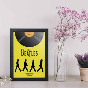 Puede incluir: Un p&oacute;ster enmarcado en blanco y negro con fondo amarillo. El p&oacute;ster presenta una silueta negra de los Beatles caminando sobre un disco. El texto "The Beatles" y "Come Together" est&aacute; impreso en el p&oacute;ster.