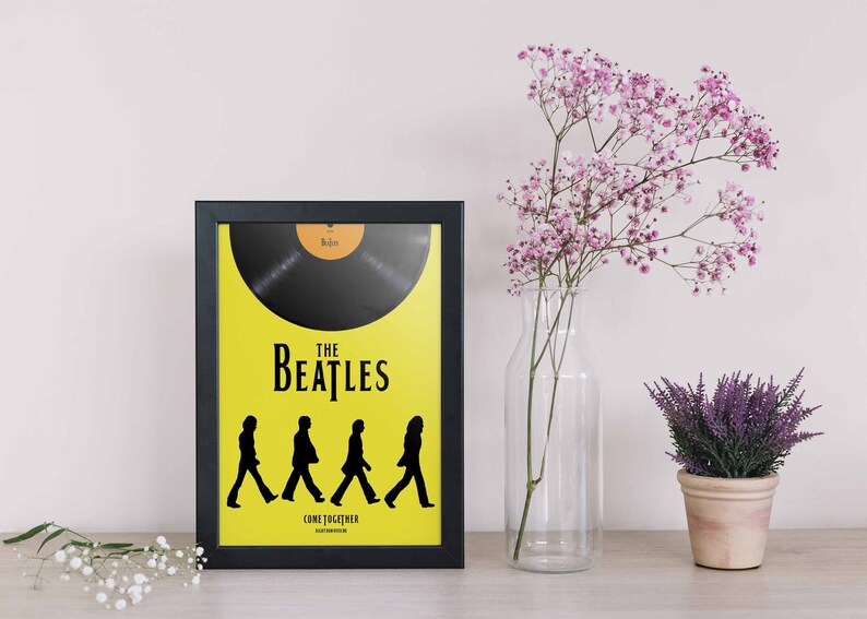 Puede incluir: Un p&oacute;ster enmarcado en blanco y negro con fondo amarillo. El p&oacute;ster presenta una silueta negra de los Beatles caminando sobre un disco. El texto "The Beatles" y "Come Together" est&aacute; impreso en el p&oacute;ster.