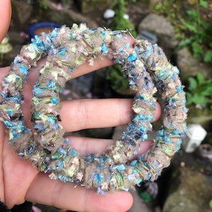 May include: A pair of large hoop earrings made of woven fabric in shades of blue, green, beige, and silver. The fabric has a fuzzy texture.