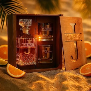 May include: A glass decanter and two matching glasses with a diamond-cut pattern, each engraved with the name JACKSON and a large letter J. The set is housed in a dark wooden box with the name JACKSON and a large letter J cut into the lid.
