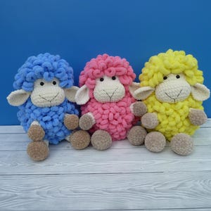 Puede incluir: Tres ovejas de peluche hechas a mano en azul, rosa y amarillo. Cada oveja tiene un cuerpo esponjoso y texturizado, cara blanca, orejas y patas marrones. Los juguetes están colocados sobre una superficie de madera azul claro y blanca.